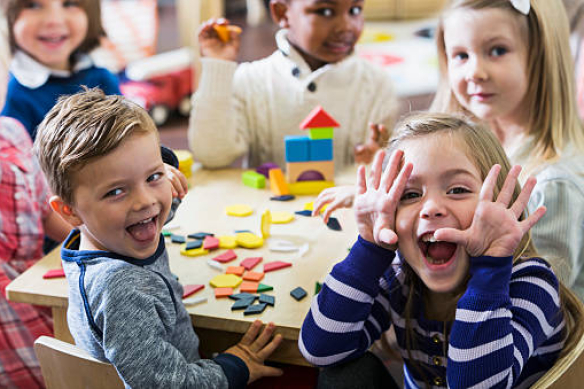 A multiracial group of preschoolers or kindergarteners having fun in the classroom.  Six children are sitting around a little wooden table playing with colorful wooden block and geometric shapes.  The playful little girl in the foreground is making a silly face at the camera.